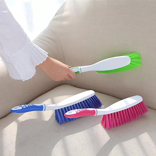 Three carpet cleaning brushes with ergonomic handles in different colors (green, blue, and pink) placed on a sofa.
