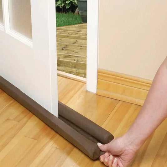 A brown under door draft stopper being installed on the bottom of a door, with a person's hand holding the stopper in place.
